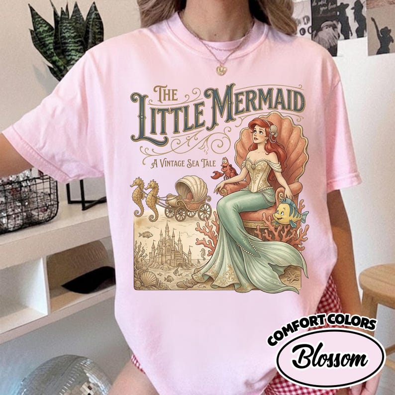 以下が含まれることがあります： 淡いピンク色のTシャツ。ヴィンテージスタイルのイラストで、リトルマーメイドが描かれています。イラストには、貝殻の玉座に座るアリエル、カニ、魚、タツノオトシゴ、馬車、お城が含まれています。「The Little Mermaid」と「A Vintage Sea Tale」の文字。