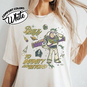 May include: A white t-shirt featuring a graphic of Buzz Lightyear saluting, surrounded by space-themed elements including a rocket, planet, and stars. The text reads "Buzz" and "To Infinity and Beyond" in a distressed, vintage-style font.