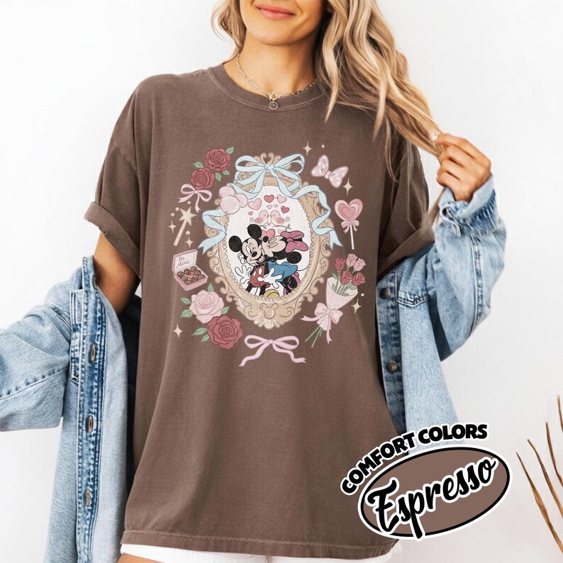 Puede incluir: Una camiseta de manga corta de color marr&oacute;n oscuro con un estampado gr&aacute;fico de Mickey y Minnie Mouse dentro de un marco ovalado ornamentado. El dise&ntilde;o incluye cintas azul claro, rosas rosas, una caja de bombones y corazones rom&aacute;nticos.