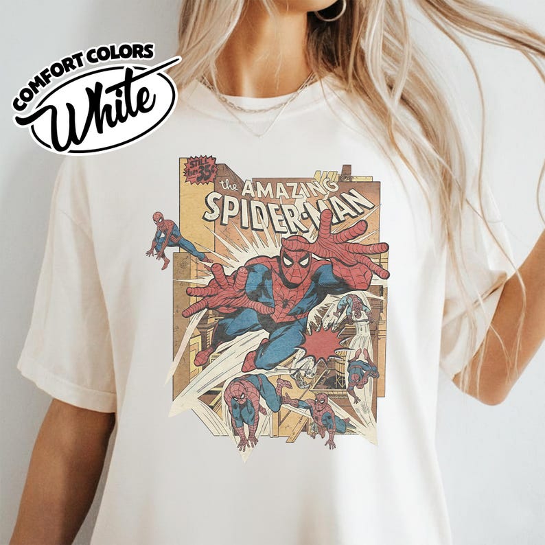K&ouml;nnte beinhalten: Wei&szlig;es T-Shirt mit einer Vintage-Comic-Illustration von Spider-Man in Aktion. Die Grafik enth&auml;lt den Text "The Amazing Spider-Man" und mehrere Bilder des Superhelden. Das Shirt hat auch das "Comfort Colors"-Logo.