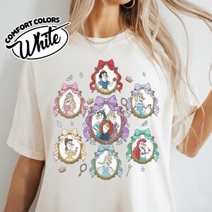 May include: White Comfort Colors t-shirt featuring a design of cartoon princesses in ornate frames with bows. The design includes Snow White, Elsa, Ariel, and other characters. The shirt has the words "Comfort Colors" and "White" in a circular design.