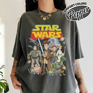 May include: Dark gray t-shirt featuring a Star Wars graphic with characters from the movie series and the text "STAR WARS" in yellow and red. The shirt also has the text "COMFORT COLORS Pepper" in a circular design.