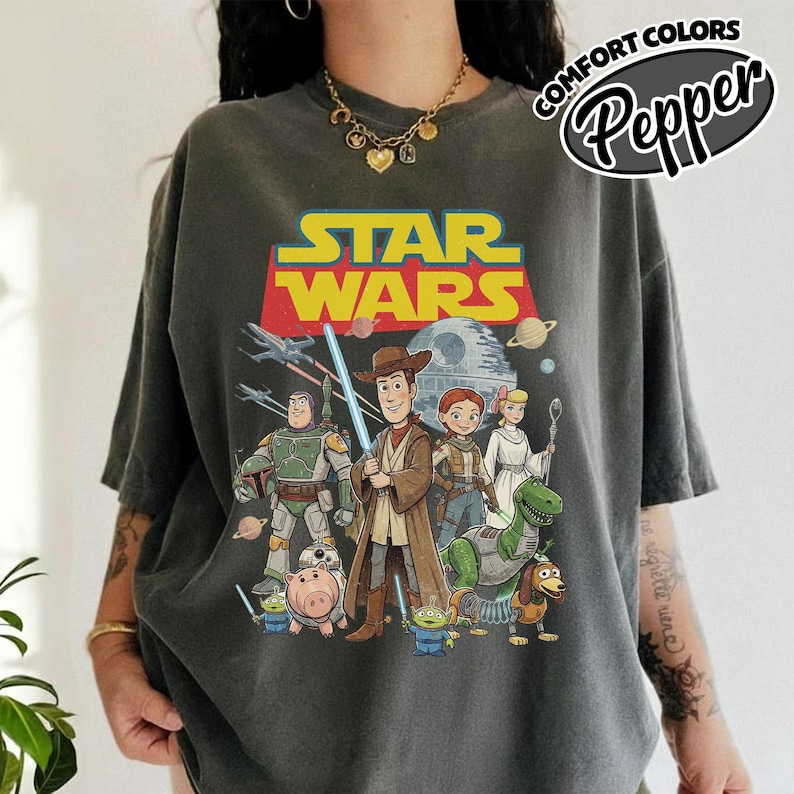 May include: Dark gray t-shirt featuring a Star Wars graphic with characters from the movie series and the text "STAR WARS" in yellow and red. The shirt also has the text "COMFORT COLORS Pepper" in a circular design.