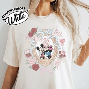 Puede incluir: Una camiseta blanca con un estampado gr&aacute;fico de Mickey y Minnie Mouse bes&aacute;ndose dentro de un marco dorado ornamentado. El dise&ntilde;o incluye rosas rojas y rosas, una cinta azul, una caja de bombones con la etiqueta Be Mine y corazones peque&ntilde;os.