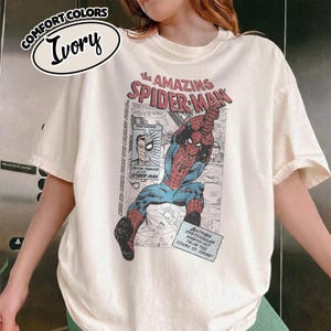 K&ouml;nnte beinhalten: Elfenbeinfarbenes T-Shirt mit einem Comic-Grafik von Spider-Man. Das Shirt hat den Text "the AMAZING SPIDER-MAN" und "COMFORT COLORS". Die Grafik enth&auml;lt den Text "PETER PARKER alias SPIDER-MAN".