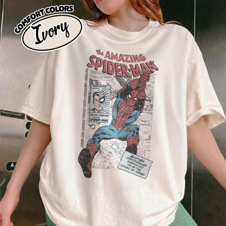 K&ouml;nnte beinhalten: Elfenbeinfarbenes T-Shirt mit einem Comic-Grafik von Spider-Man. Das Shirt hat den Text "the AMAZING SPIDER-MAN" und "COMFORT COLORS". Die Grafik enth&auml;lt den Text "PETER PARKER alias SPIDER-MAN".