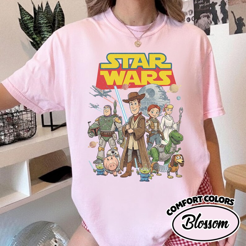 May include: A light pink t-shirt featuring a Star Wars graphic with various characters and the text "STAR WARS" in yellow and blue. The shirt also has the text "COMFORT COLORS" and "Blossom".