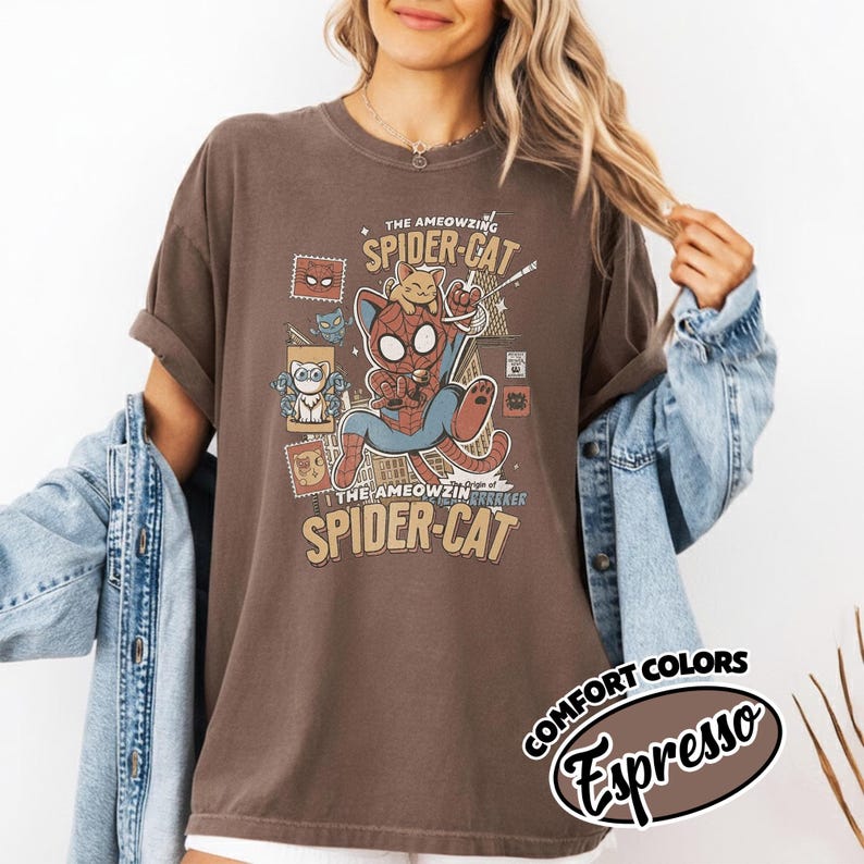 May include: Brown t-shirt featuring a cartoon Spider-Cat design. The graphic includes a cat in a Spider-Man suit, the words "The AMEOWZING SPIDER-CAT" and "COMFORT COLORS Espresso". The shirt has rolled-up sleeves.