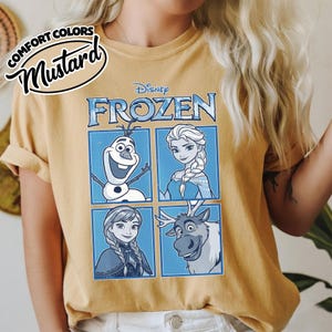 May include: Mustard-colored t-shirt featuring a Disney Frozen graphic. The graphic includes images of Olaf, Elsa, Anna, and Sven, set against a blue background. The shirt has rolled-up sleeves and the words "Comfort Colors" and "Mustard" are printed on the top left.