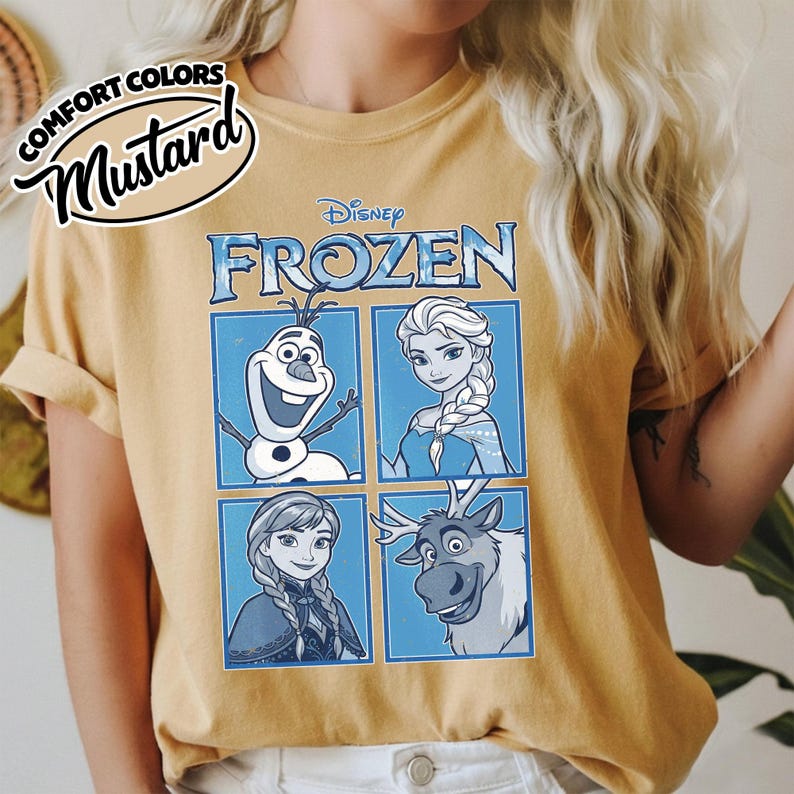 May include: Mustard-colored t-shirt featuring a Disney Frozen graphic. The graphic includes images of Olaf, Elsa, Anna, and Sven, set against a blue background. The shirt has rolled-up sleeves and the words "Comfort Colors" and "Mustard" are printed on the top left.