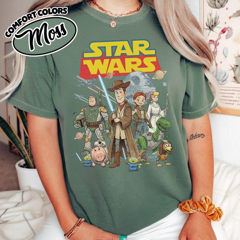 May include: Moss green t-shirt featuring a mashup of Star Wars and Toy Story characters. The design includes Woody as a Jedi, Buzz Lightyear, and other characters from both franchises. The words "STAR WARS" are in yellow and red.