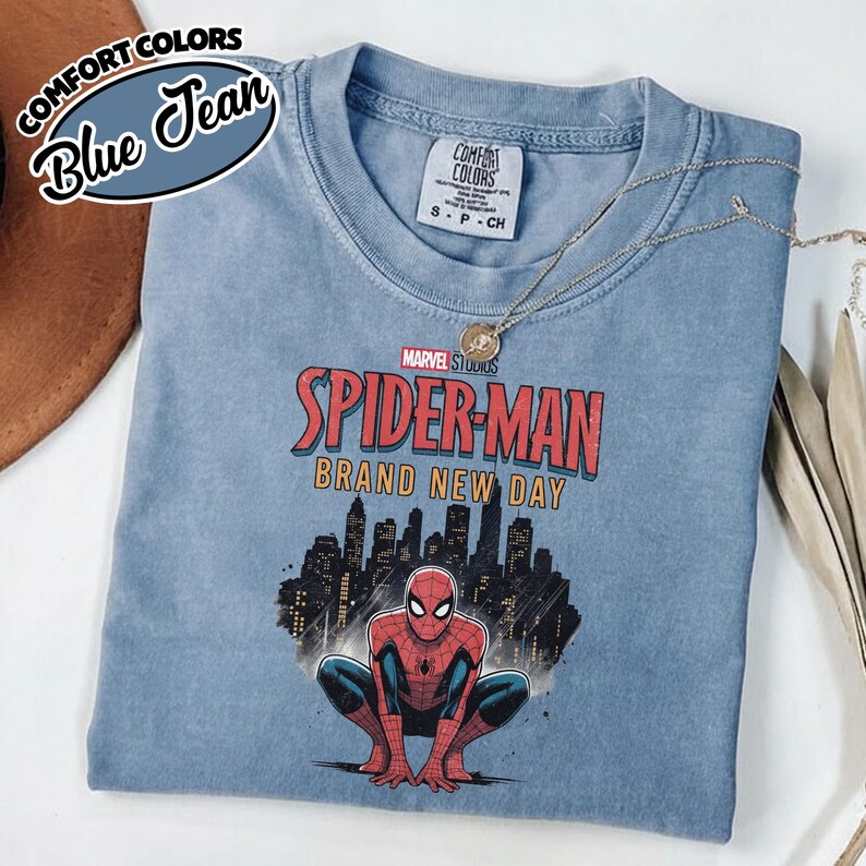 Puede incluir: Una camiseta de manga corta color azul jean con un gr&aacute;fico de Spider-Man agachado frente al horizonte de una ciudad. El texto dice Marvel Studios Spider-Man Brand New Day. La camiseta tiene cuello redondo y una etiqueta de marca Comfort Colors.