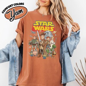 May include: Rust-colored t-shirt featuring a graphic of Star Wars characters, including Woody, Buzz Lightyear, and others. The Star Wars logo is displayed above the characters. The shirt also has the text "Comfort Colors" and "Yam".