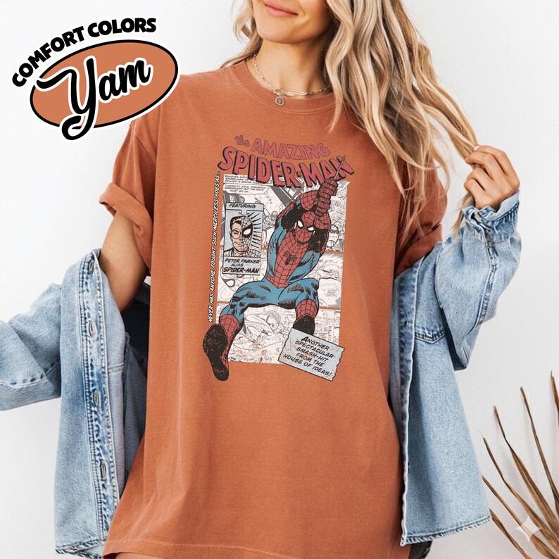 K&ouml;nnte beinhalten: Ein rostfarbenes T-Shirt mit einer Vintage-Comic-Grafik von Spider-Man. Das Design enth&auml;lt den Text "The Amazing Spider-Man" und andere Comic-Elemente. Das Shirt hat hochgekrempelte &Auml;rmel und eine lockere Passform.