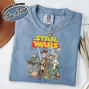 May include: A blue Comfort Colors t-shirt featuring a Star Wars and Toy Story mashup graphic. The graphic includes characters like Woody, Buzz Lightyear, and Darth Vader. The shirt has the text "STAR WARS" in yellow and red.