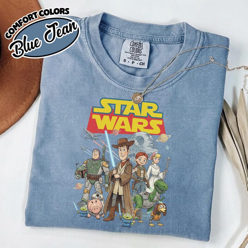 May include: A blue Comfort Colors t-shirt featuring a Star Wars and Toy Story mashup graphic. The graphic includes characters like Woody, Buzz Lightyear, and Darth Vader. The shirt has the text "STAR WARS" in yellow and red.