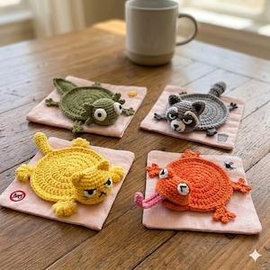 May include: Four handmade crochet coasters in the shape of animals: a green lizard, a gray raccoon, a yellow cat, and an orange frog. Each coaster is on a pink square. A white mug sits in the background.