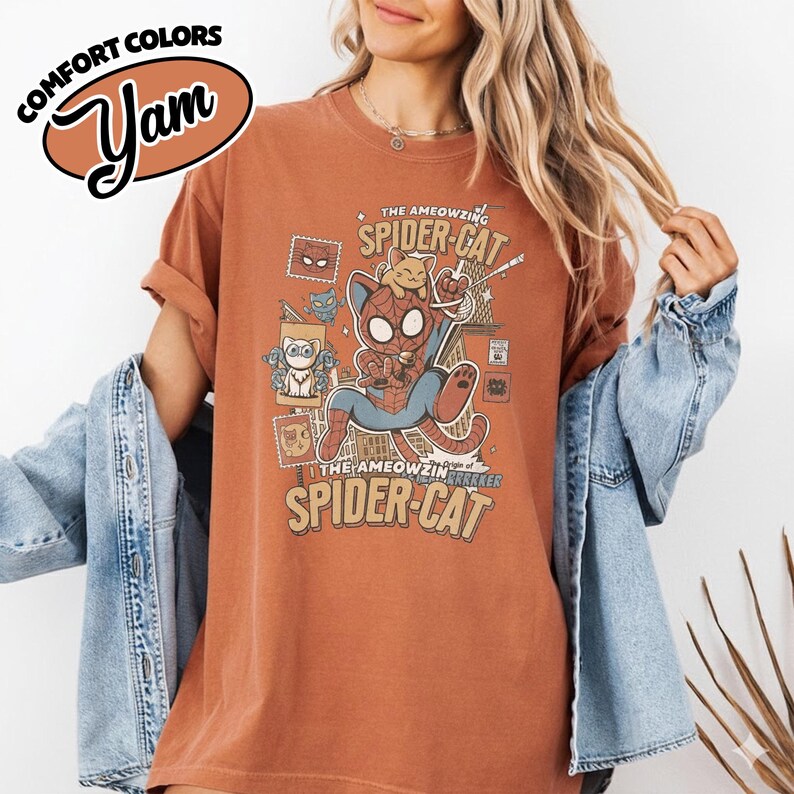 May include: Rust-colored t-shirt featuring a cartoon Spider-Cat design. The graphic includes a cat dressed as Spider-Man, with the text "The AMEOWZING SPIDER-CAT" and "The AMEOWZING PARKER SPIDER-CAT". The shirt has rolled-up sleeves.