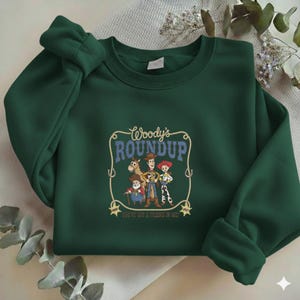 Puede incluir: Una sudadera de cuello redondo verde oscuro con un estampado gr&aacute;fico de Woody, Jessie, Bullseye y Stinky Pete de Toy Story. El texto dice Woody's ROUNDUP y YOU'VE GOT A FRIEND IN ME, enmarcado por un dise&ntilde;o de lazo con dos estrellas de sheriff.