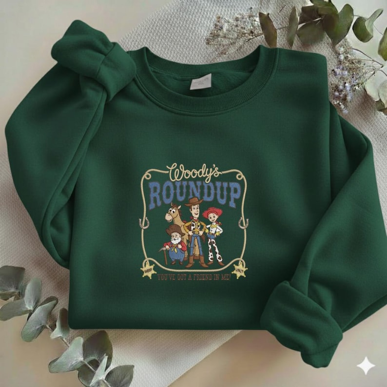 Puede incluir: Una sudadera de cuello redondo verde oscuro con un estampado gr&aacute;fico de Woody, Jessie, Bullseye y Stinky Pete de Toy Story. El texto dice Woody's ROUNDUP y YOU'VE GOT A FRIEND IN ME, enmarcado por un dise&ntilde;o de lazo con dos estrellas de sheriff.