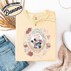 Puede incluir: Una camiseta de color amarillo p&aacute;lido con un gr&aacute;fico de Mickey y Minnie Mouse bes&aacute;ndose dentro de un marco ovalado ornamentado. El dise&ntilde;o incluye rosas rojas y rosas, una cinta azul, una caja de bombones con la etiqueta Be Mine y una piruleta en forma de coraz&oacute;n.