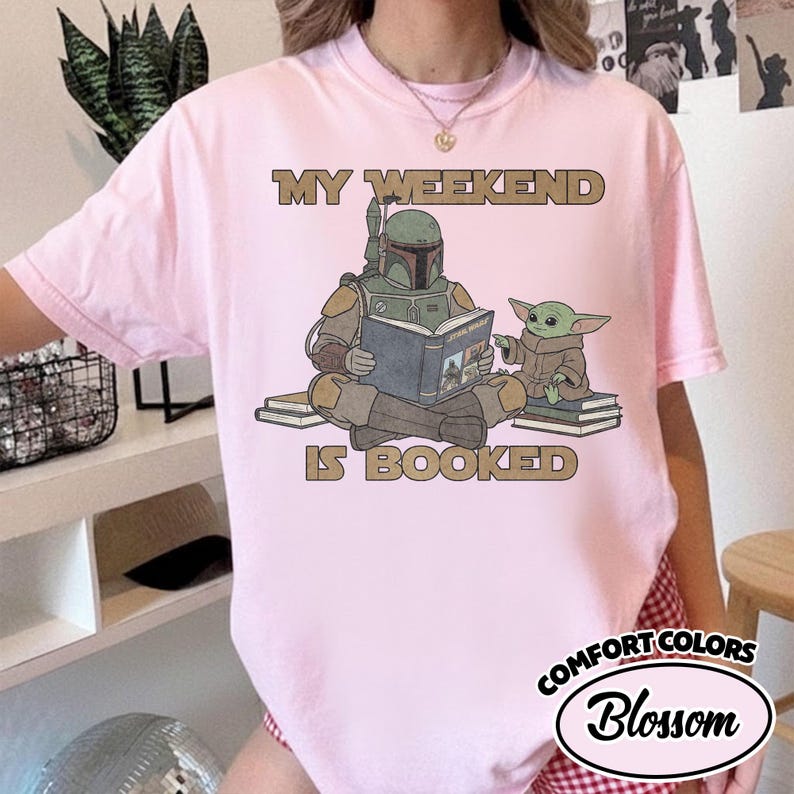 Może przedstawiać: Jasnor&oacute;żowa koszulka z napisem "MY WEEKEND IS BOOKED" nad ilustracją Boba Fetta i Grogu czytających książki. Koszulka ma etykietę Comfort Colors Blossom.