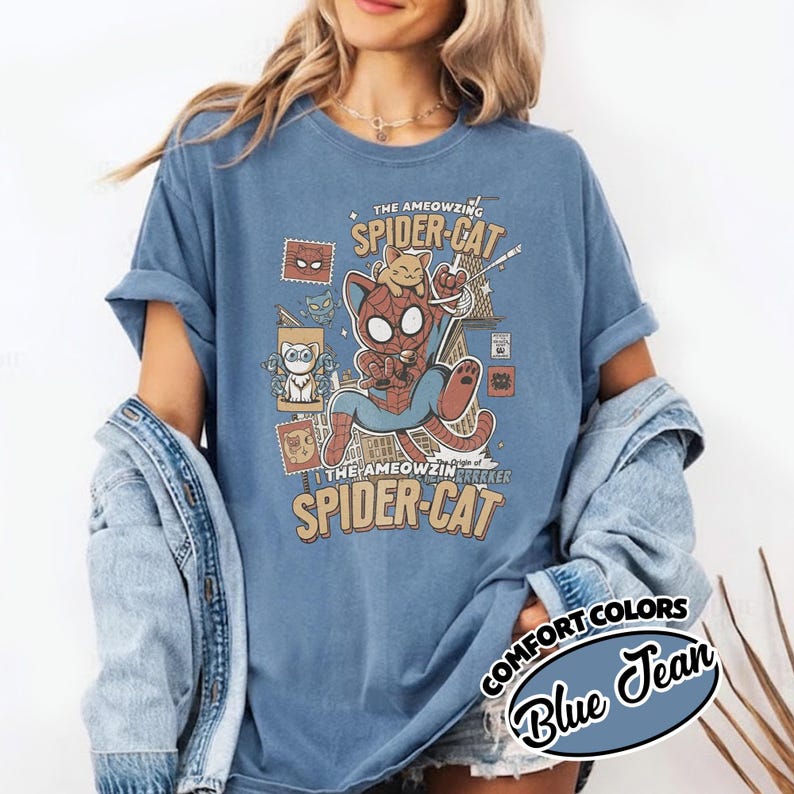May include: Blue jean colored t-shirt with a graphic of Spider-Man as a cat, labeled "The Ameowzing Spider-Cat." The design includes stamps and the text "Comfort Colors." The shirt is a relaxed fit.