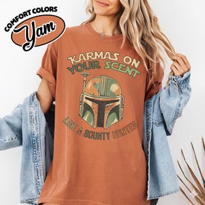 May include: A rust-colored t-shirt with the text "Karma's on Your Scent Like a Bounty Hunter" and a graphic of a Mandalorian helmet. The shirt has rolled-up sleeves and a relaxed fit. The brand name "Yam" is in a brown oval.