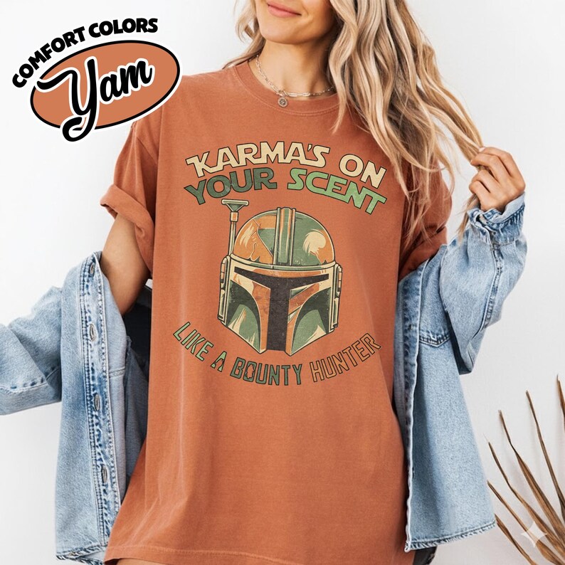 May include: A rust-colored t-shirt with the text "Karma's on Your Scent Like a Bounty Hunter" and a graphic of a Mandalorian helmet. The shirt has rolled-up sleeves and a relaxed fit. The brand name "Yam" is in a brown oval.