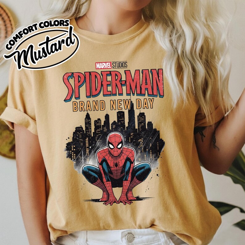 Puede incluir: Una camiseta de manga corta color amarillo mostaza con un gr&aacute;fico de Spider-Man agachado frente a un horizonte urbano negro. El texto dice Marvel Studios Spider-Man Brand New Day. Una etiqueta circular en la esquina dice Comfort Colors Mustard.