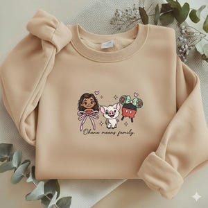May include: A beige crewneck sweatshirt featuring a central graphic of a cartoon character with long brown hair, a small pig, and a Mickey Mouse-shaped treat with a bow. The text "Ohana means family." is printed in black cursive below the characters.