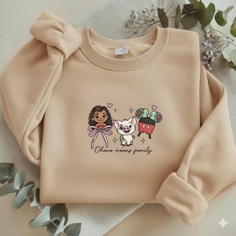 May include: A beige crewneck sweatshirt featuring a central graphic of a cartoon character with long brown hair, a small pig, and a Mickey Mouse-shaped treat with a bow. The text "Ohana means family." is printed in black cursive below the characters.