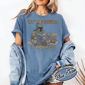 Może przedstawiać: Niebieska koszulka Comfort Colors z napisem "MY WEEKEND IS BOOKED". Grafika przedstawia postać w zielonej zbroi czytającą książkę z małą zieloną istotą. Koszulka jest w kolorze blue jean.