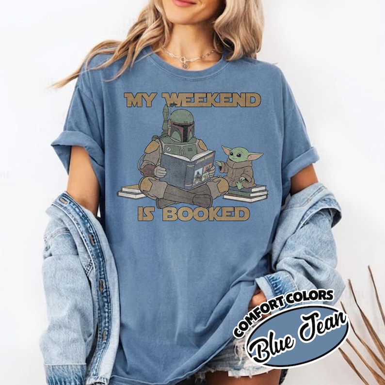 Może przedstawiać: Niebieska koszulka Comfort Colors z napisem "MY WEEKEND IS BOOKED". Grafika przedstawia postać w zielonej zbroi czytającą książkę z małą zieloną istotą. Koszulka jest w kolorze blue jean.