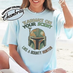 May include: Light blue t-shirt with the text "Karma's on Your Scent Like a Bounty Hunter" and a graphic of a Mandalorian helmet in shades of green, brown, and orange. The shirt has the "Comfort Colors" and "Chambray" logos.