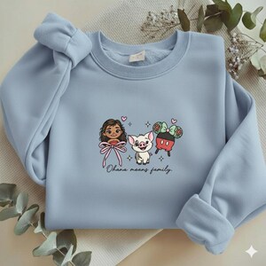 May include: A light blue crewneck sweatshirt featuring a central graphic of a cartoon girl, a piglet, and a pair of mouse ears with a bow. The text "Ohana means family." is printed below the characters, accompanied by small heart and star icons.