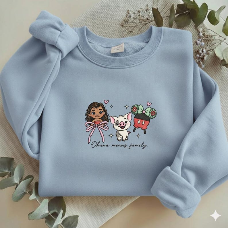 May include: A light blue crewneck sweatshirt featuring a central graphic of a cartoon girl, a piglet, and a pair of mouse ears with a bow. The text "Ohana means family." is printed below the characters, accompanied by small heart and star icons.