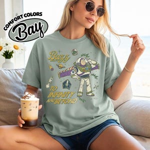 May include: A sage green t-shirt featuring a graphic of the character Buzz Lightyear saluting, surrounded by space-themed illustrations including a rocket, planet, and stars, with the text Buzz To Infinity And Beyond printed in a vintage style.