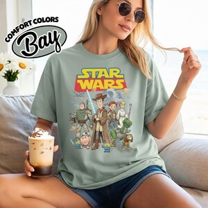May include: Sage green t-shirt featuring a Star Wars graphic with characters from the Toy Story franchise. The shirt has the words "STAR WARS" in yellow and red. The shirt also has the words "COMFORT COLORS" and "Bay" in a white oval.