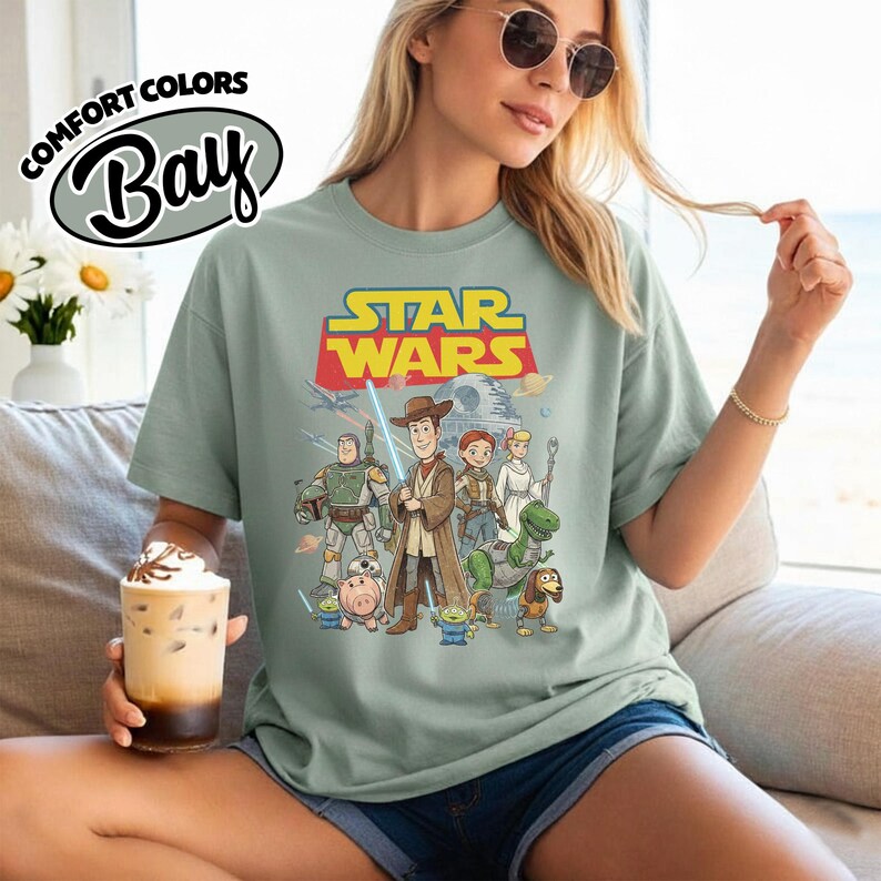 May include: Sage green t-shirt featuring a Star Wars graphic with characters from the Toy Story franchise. The shirt has the words "STAR WARS" in yellow and red. The shirt also has the words "COMFORT COLORS" and "Bay" in a white oval.