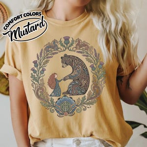 May include: Mustard-colored t-shirt with a circular graphic featuring a bear and a person holding hands, surrounded by a floral wreath. The bear has intricate tribal-style patterns. The text "COMFORT COLORS" and "Mustard" are in the upper left corner.