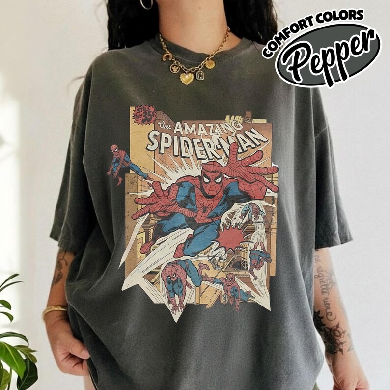 K&ouml;nnte beinhalten: Dunkelgraues T-Shirt mit einem Vintage-Comic-Grafik von Spider-Man. Das Design enth&auml;lt den Text "The Amazing Spider-Man" und die Worte "Comfort Colors Pepper" in einem kreisf&ouml;rmigen Logo.