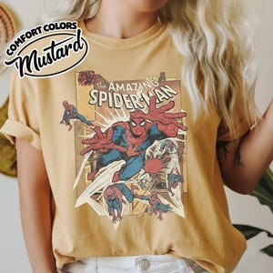 K&ouml;nnte beinhalten: Senffarbenes T-Shirt mit einem Vintage-Comic-Cover von Spider-Man. Die Grafik zeigt Spider-Man in Aktion mit dem Text "The Amazing Spider-Man". Das Shirt hat hochgekrempelte &Auml;rmel.