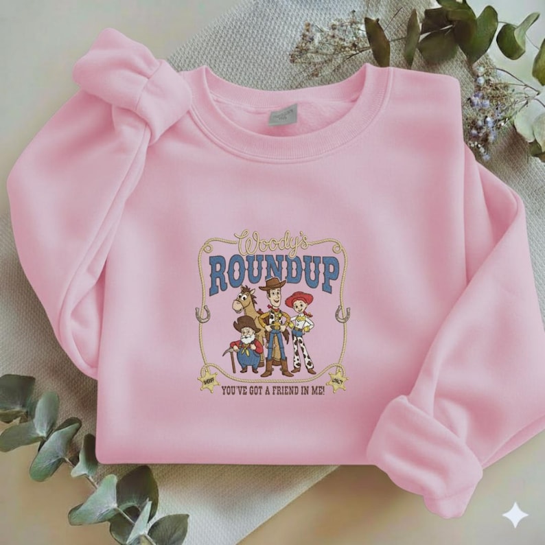 Puede incluir: Una sudadera de cuello redondo rosa claro con un estampado gr&aacute;fico de Woody, Jessie, Bullseye y Stinky Pete de Toy Story. El texto dice Woody's ROUNDUP y YOU'VE GOT A FRIEND IN ME! El dise&ntilde;o est&aacute; enmarcado por un borde de cuerda con estrellas.