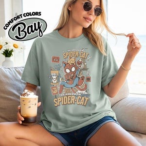 May include: A sage green t-shirt with a graphic of a cat dressed as Spider-Man, with the text "The AMEOWZING SPIDER-CAT". The shirt also has the text "COMFORT COLORS Bay" in a white oval. The shirt is worn by a person.
