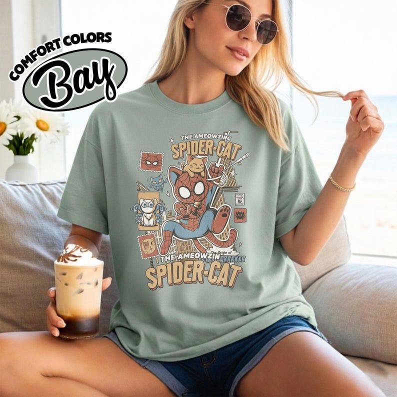 May include: A sage green t-shirt with a graphic of a cat dressed as Spider-Man, with the text "The AMEOWZING SPIDER-CAT". The shirt also has the text "COMFORT COLORS Bay" in a white oval. The shirt is worn by a person.