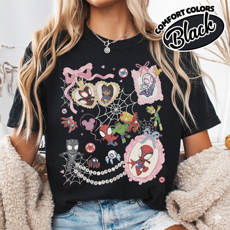 K&ouml;nnte beinhalten: Schwarzes T-Shirt mit einer farbenfrohen Collage von Spider-Man-Charakteren in rosa Rahmen und Spinnennetz-Designs. Das Shirt hat den Text "COMFORT COLORS Black" in einem kreisf&ouml;rmigen Design. Das Design umfasst verschiedene Charaktere und dekorative Elemente.