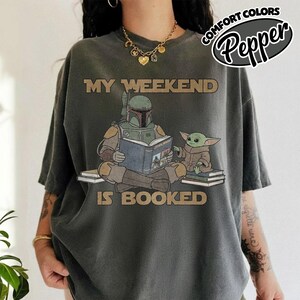 Może przedstawiać: Ciemnoszara koszulka z napisem "MY WEEKEND IS BOOKED" nad ilustracją Boba Fetta czytającego książkę Grogu. Koszulka ma luźny kr&oacute;j i logo "Comfort Colors" w prawym g&oacute;rnym rogu.