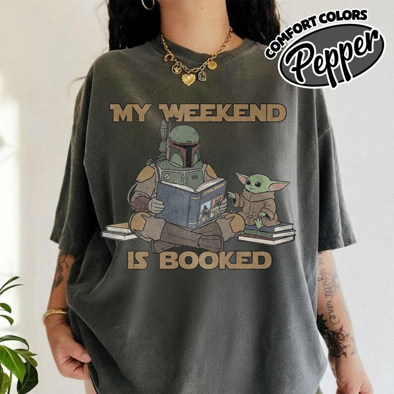 Może przedstawiać: Ciemnoszara koszulka z napisem "MY WEEKEND IS BOOKED" nad ilustracją Boba Fetta czytającego książkę Grogu. Koszulka ma luźny kr&oacute;j i logo "Comfort Colors" w prawym g&oacute;rnym rogu.