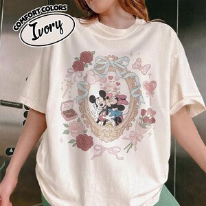 Puede incluir: Una camiseta de color marfil con un gr&aacute;fico de Mickey y Minnie Mouse bes&aacute;ndose dentro de un marco ovalado ornamentado. El dise&ntilde;o incluye rosas rojas, una cinta azul, una caja de bombones con la etiqueta Be Mine, una piruleta y un ramo.